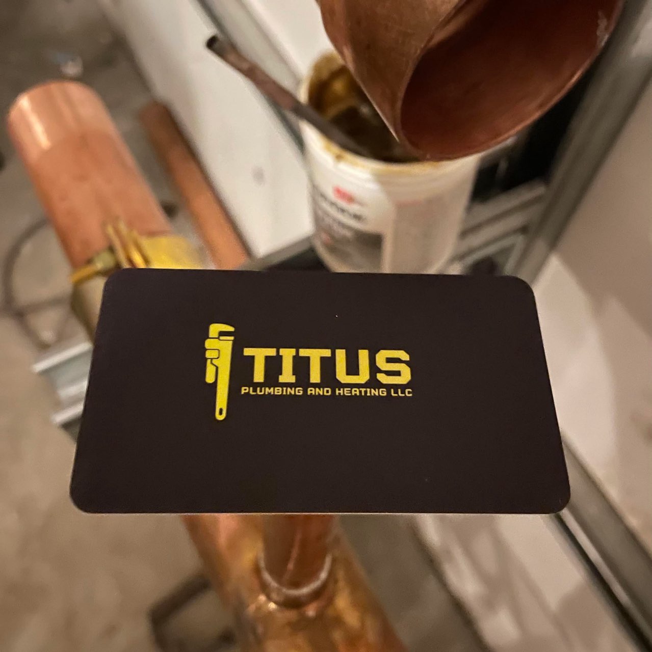 Titus Plumbing and Heating LLC business card displayed near copper pipes, representing plumbing and heating services.